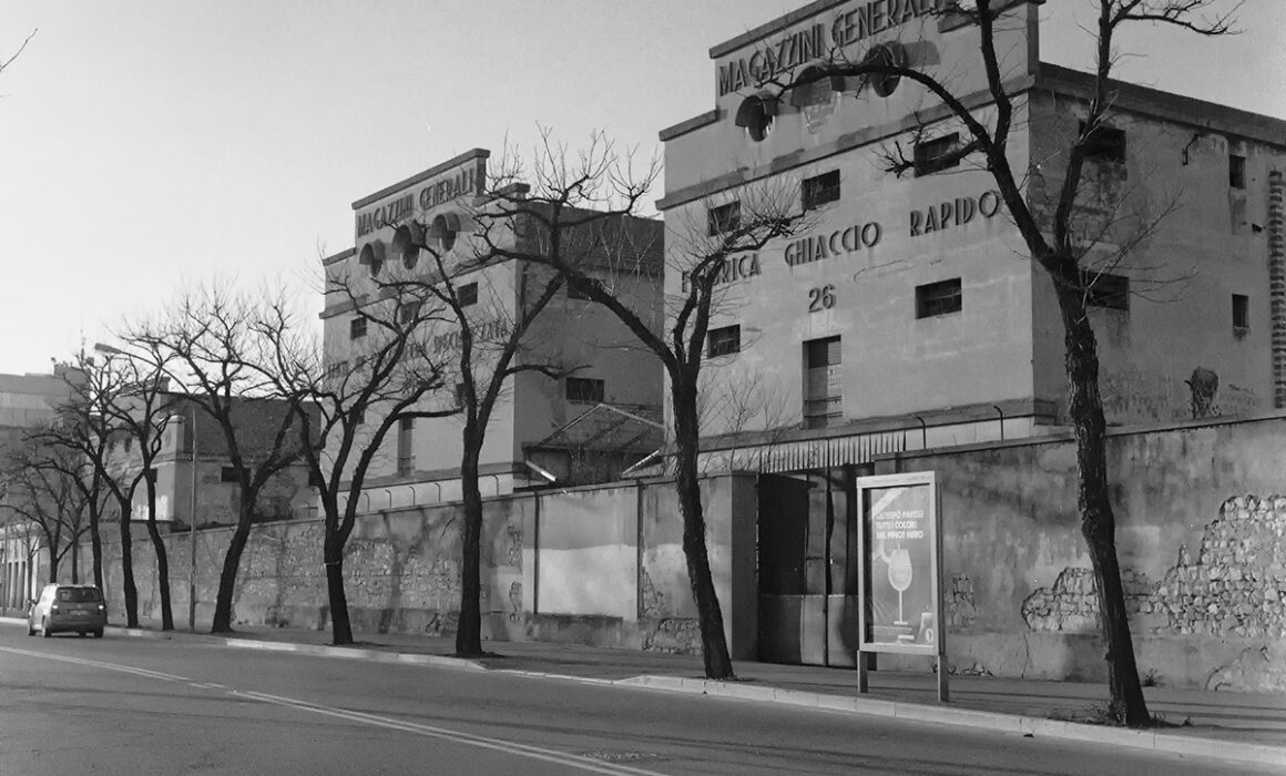 Two industrial buildings labeled “Magazzini Generali” and “Fabbrica Ghiaccio Rapido” along a street with bare trees.
